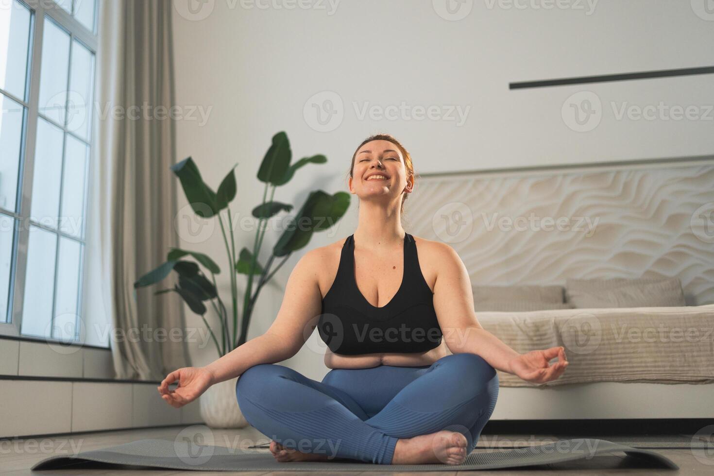 Yoga mindfulness meditation. Woman practicing yoga in living room at home. Plus size female sitting in lotus pose meditating smiling relaxing indoor. Girl doing breathing practice. Yoga at home photo