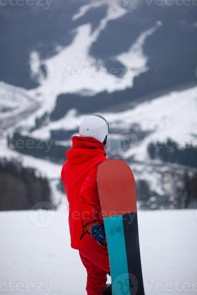 A man in a red jacket with a snowboard stands on a mountainside. photo