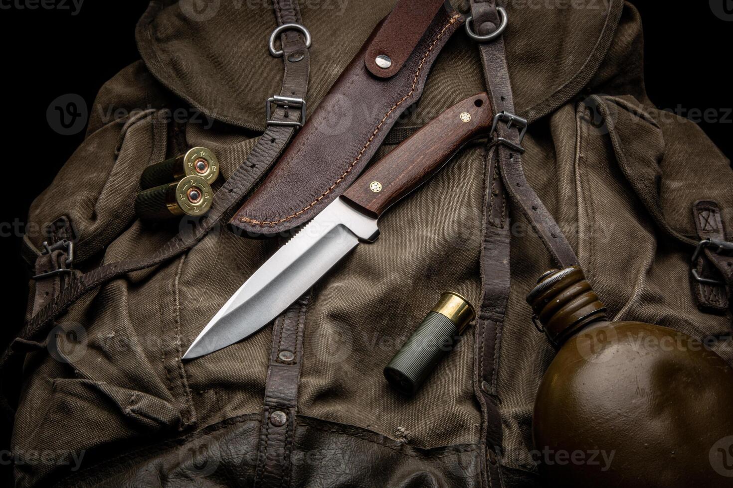 Hunting knife with a wooden handle on a vintage canvas backpack. 12-caliber shotgun shells and a water canteen are nearby. Hunter's set. Dark back. photo