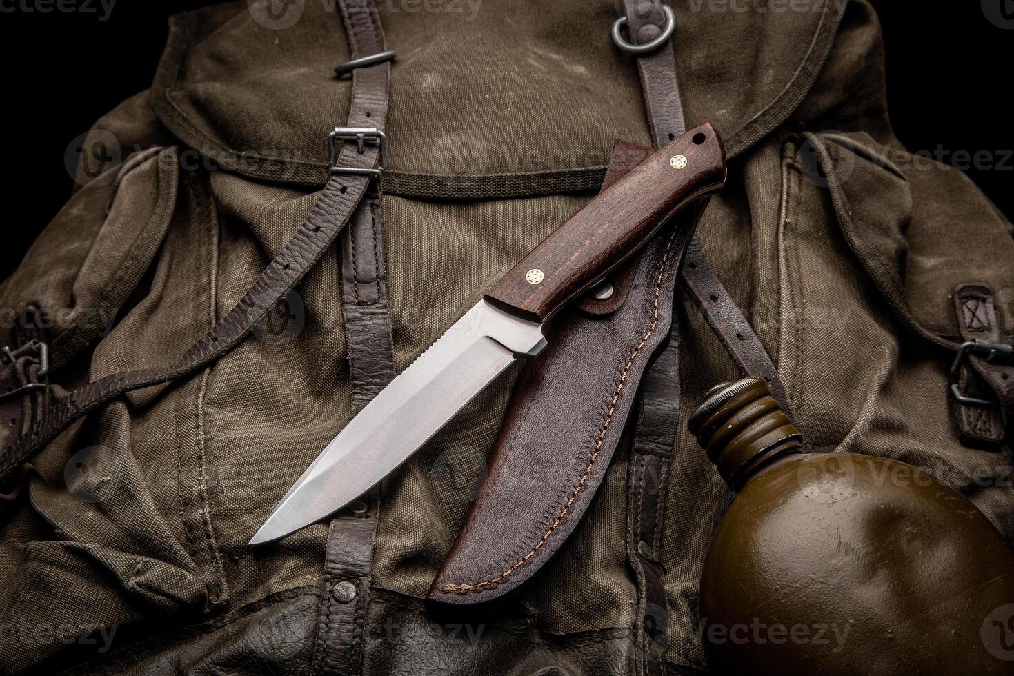 A hunting knife with a wooden handle and a leather sheath on a khaki canvas backpack. Weapons for self-defense and survival. Dark back. photo