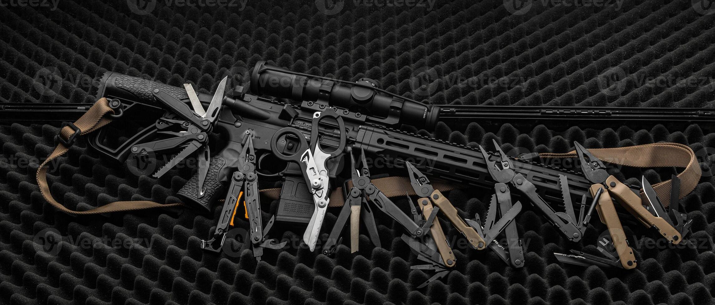 Various multi-tools lie near the automatic carbine on a gray foam surface. Dark back. photo