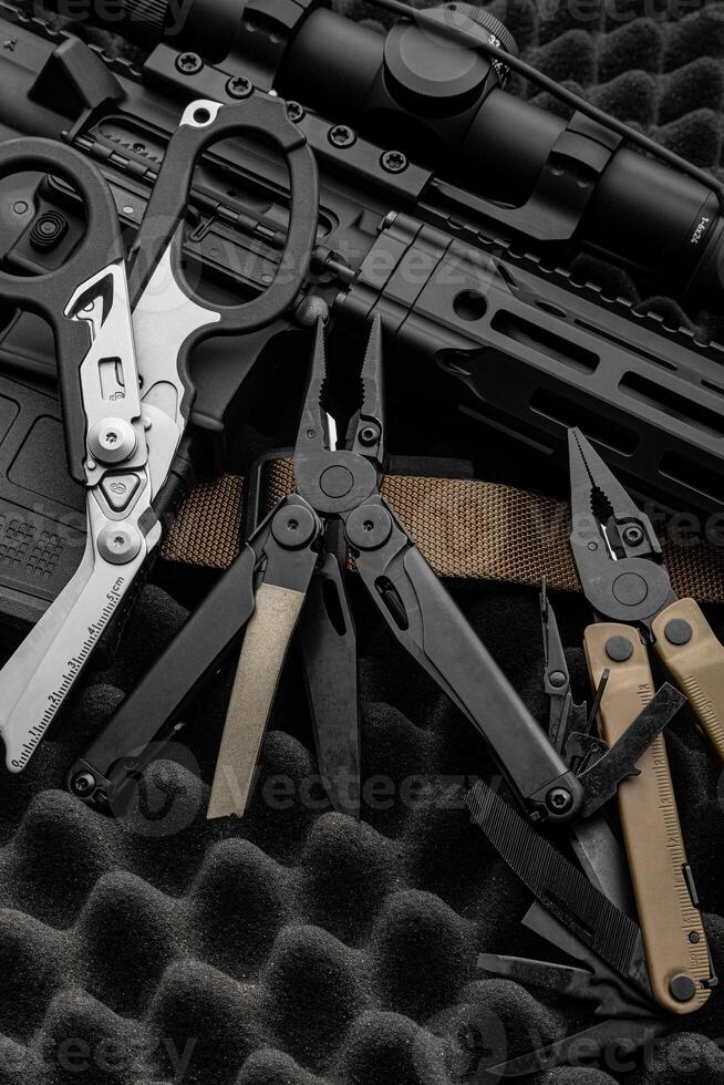 Modern multitool with many tools. A portable multi-tasking military tool lies next to the weapon. Multifunctional scissors. Dark back. photo