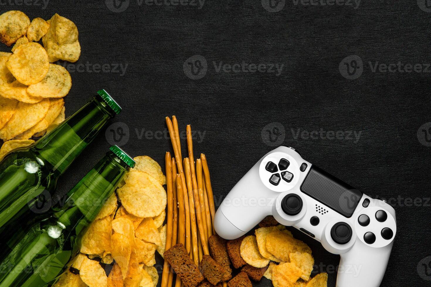 Modern white gamepad among chips, crackers and bottles of beer on a black. Game concept. photo