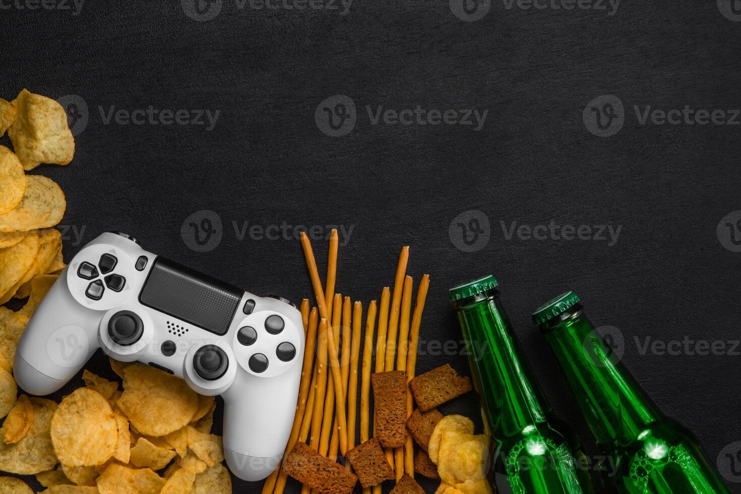 Modern white gamepad among chips, crackers and bottles of beer on a black. Game concept. photo