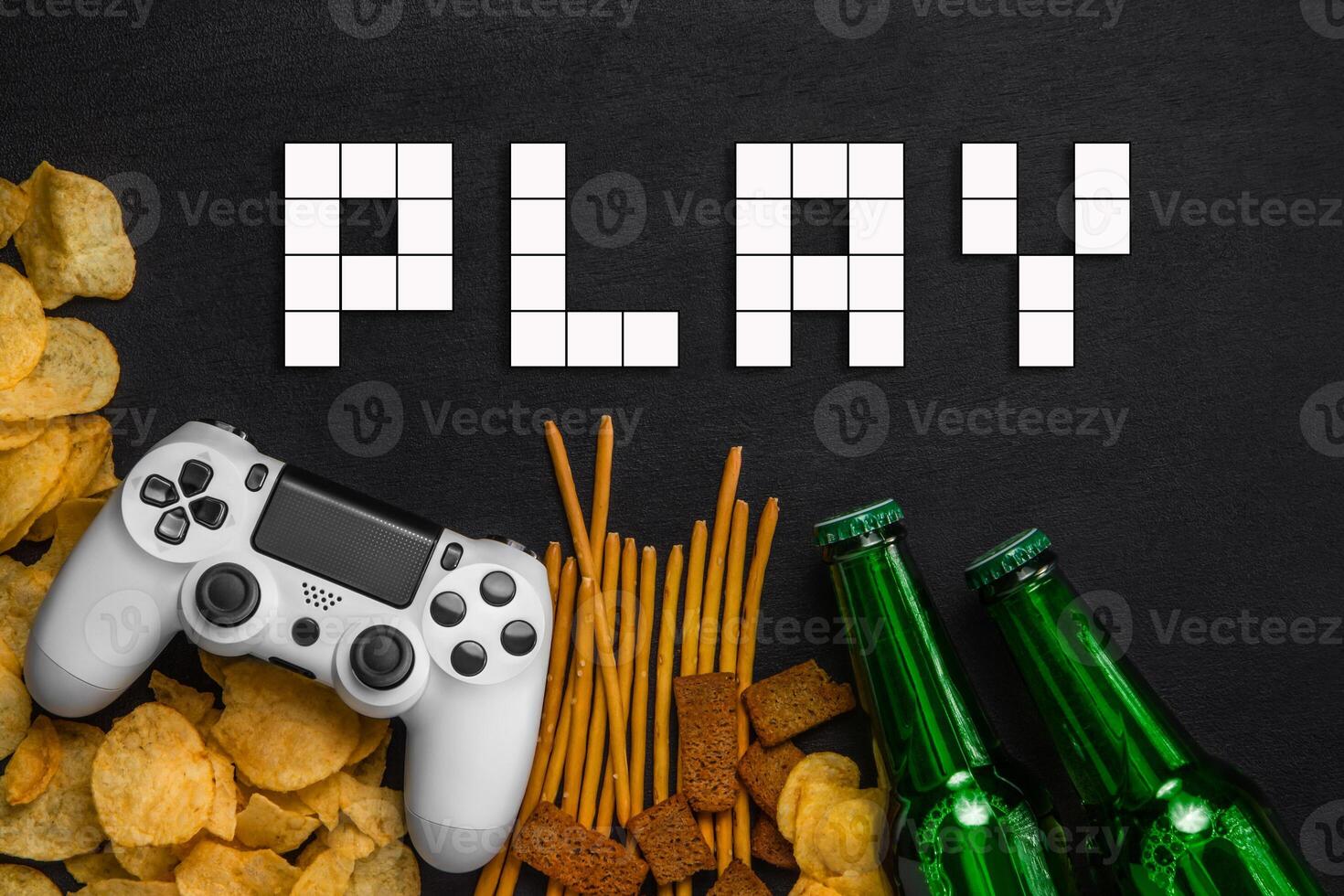 Modern white gamepad among chips, crackers and bottles of beer on a black. Game concept. photo