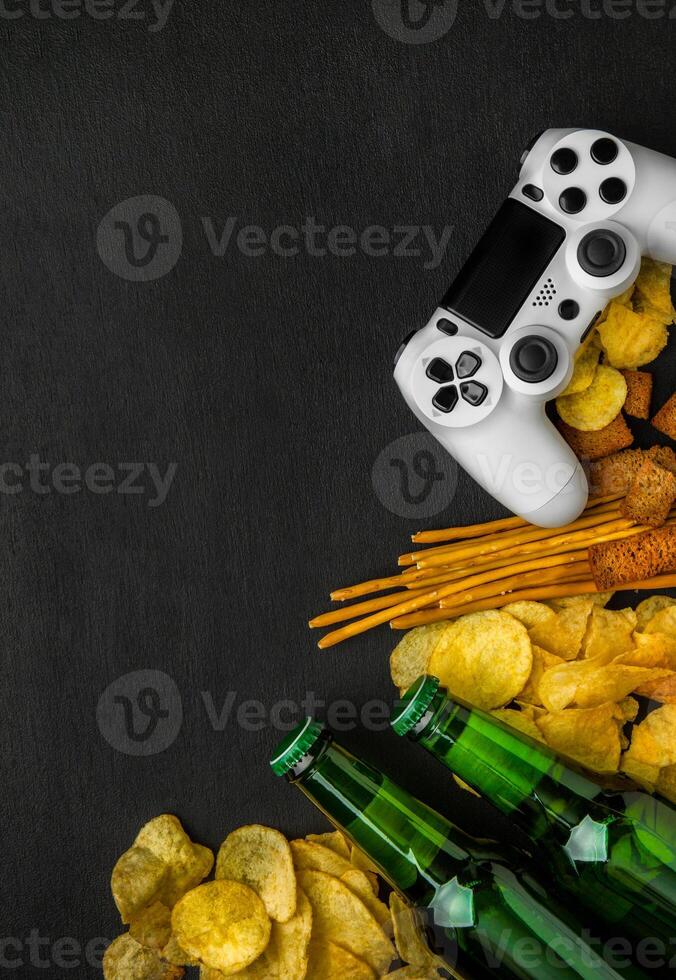 Modern white gamepad among chips, crackers and bottles of beer on a black. Game concept. photo