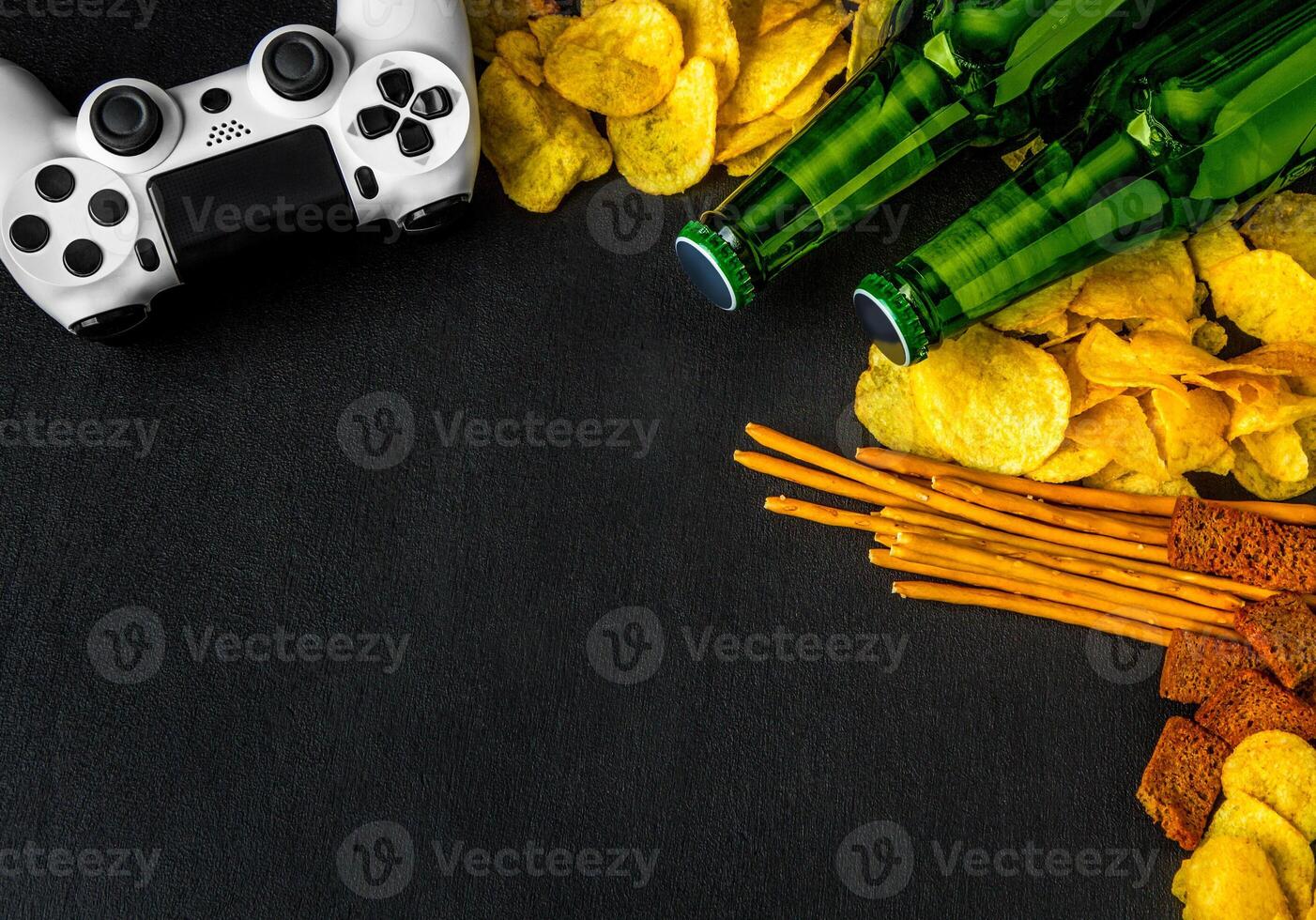 Modern white gamepad among chips, crackers and bottles of beer on a black. Game concept. photo