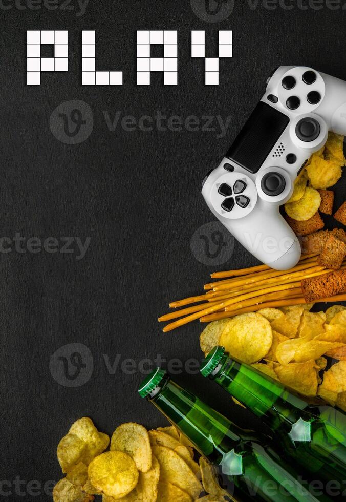 Modern white gamepad among chips, crackers and bottles of beer on a black. Game concept. photo