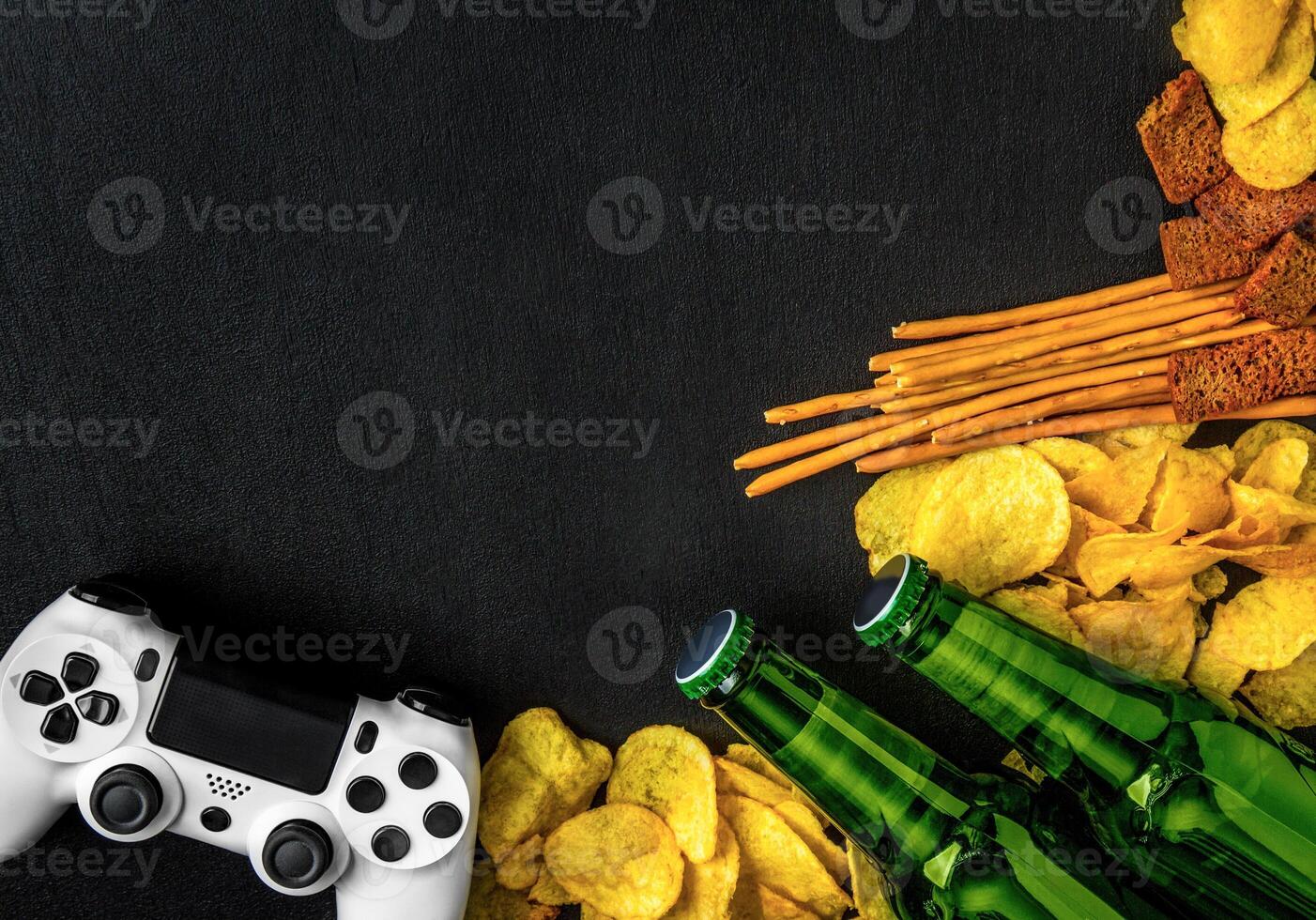 Modern white gamepad among chips, crackers and bottles of beer on a black. Game concept. photo