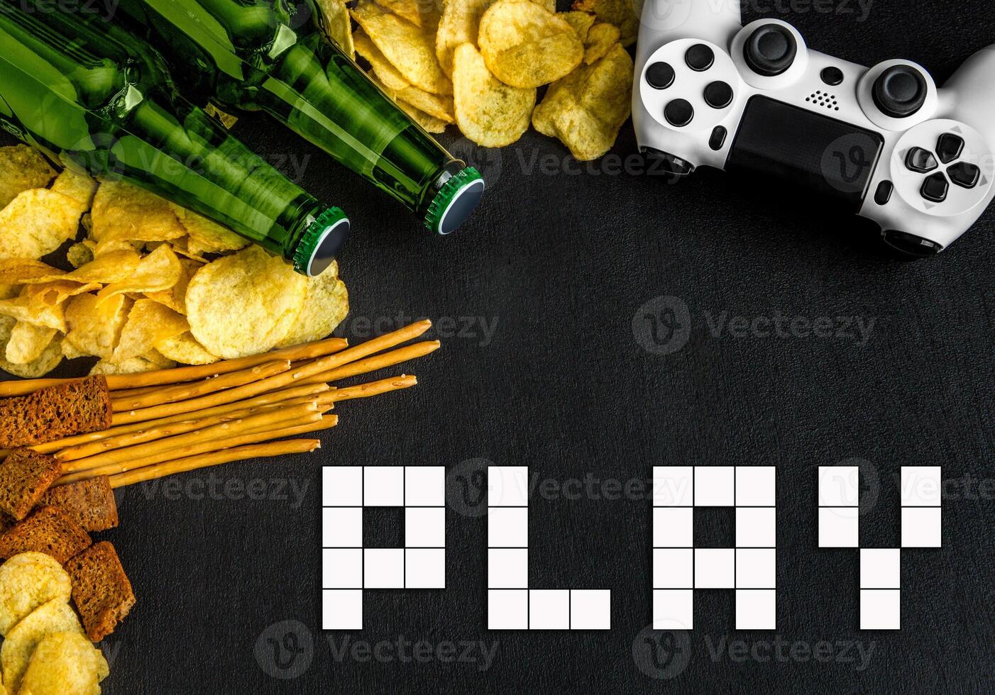 Modern white gamepad among chips, crackers and bottles of beer on a black. Game concept. photo