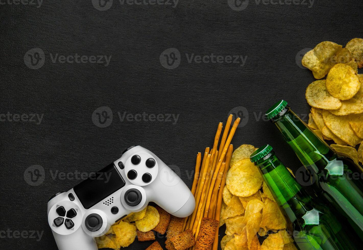Modern white gamepad among chips, crackers and bottles of beer on a black. Game concept. photo