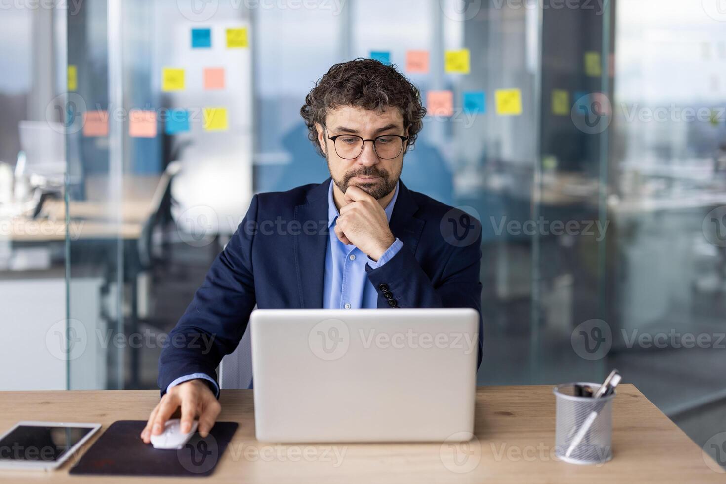 Businessman in suit working on laptop at modern office. Professional focused on tasks with sticky notes in background indicating organization and planning. Ideal for business corporate photo