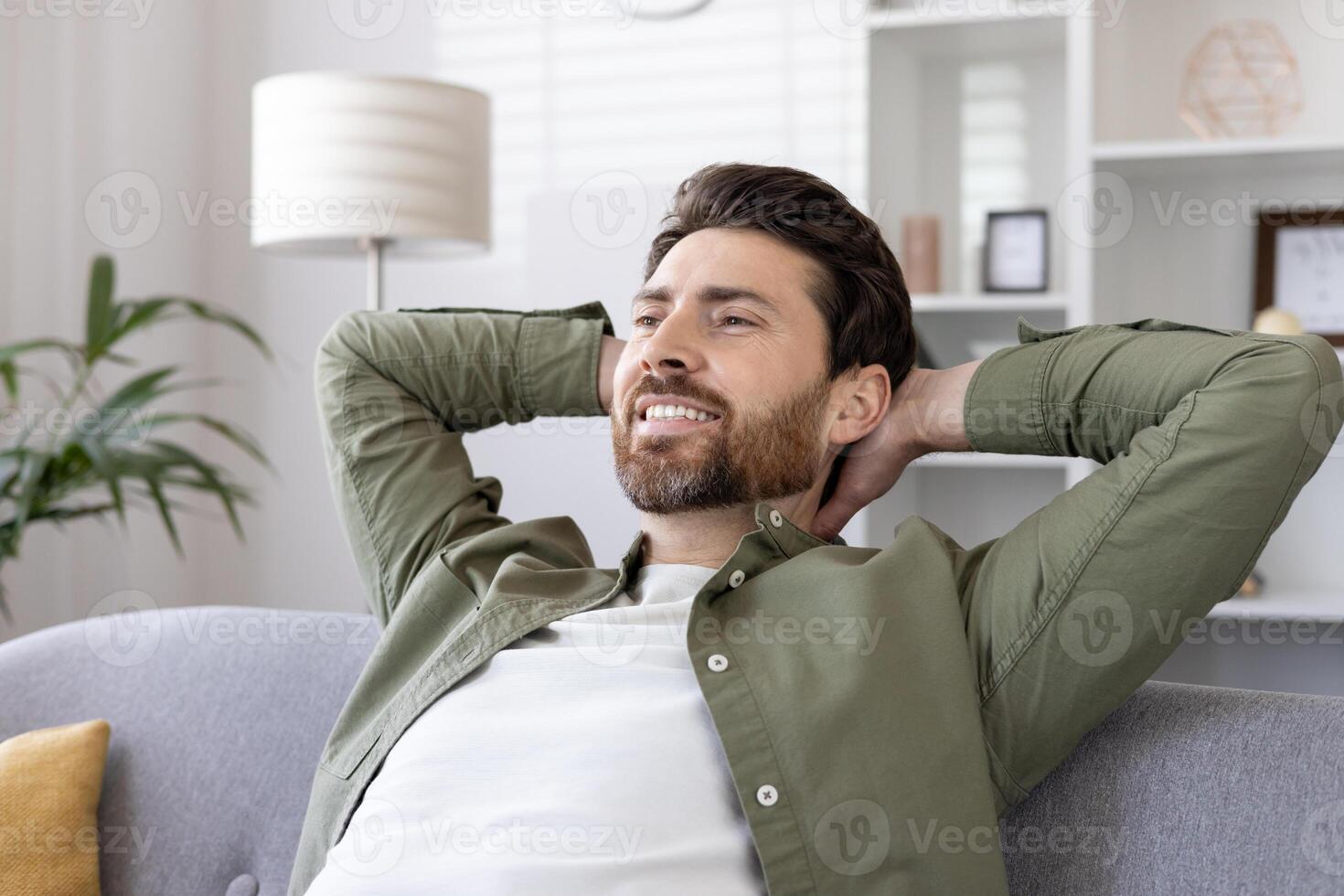 Relaxed man sitting on sofa with hands behind head, enjoying leisure time at home. Man smiling while resting and feeling stress-free indoors. photo