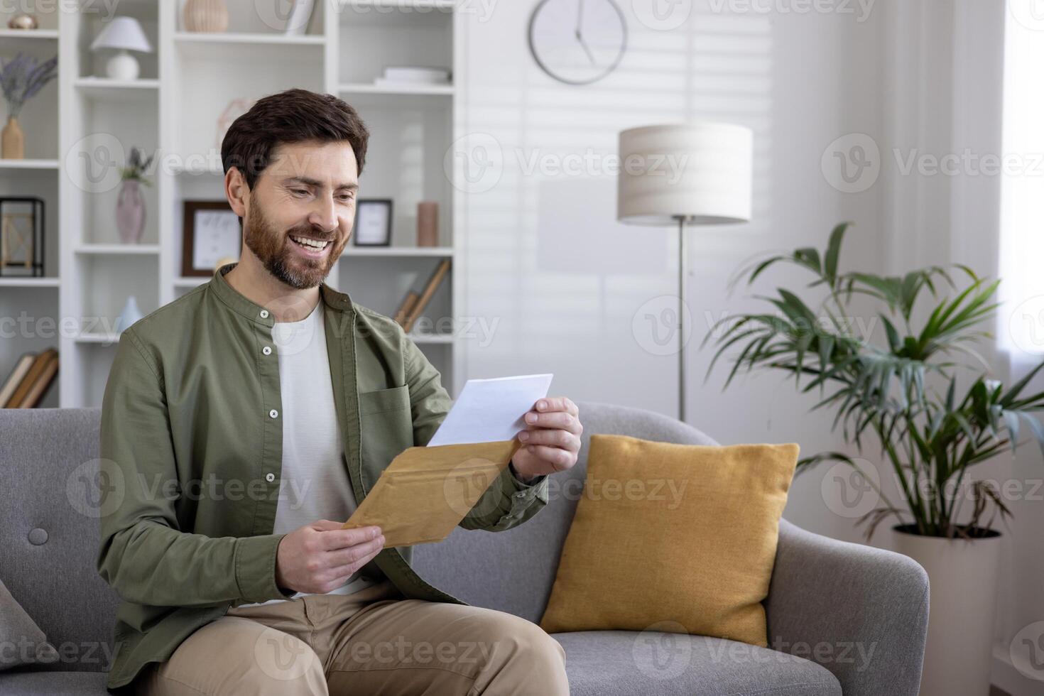 Smiling man reading letter while sitting on couch in modern living room. Happy expressions, casual attire, cozy environment conveying messages of joy and relaxation. photo