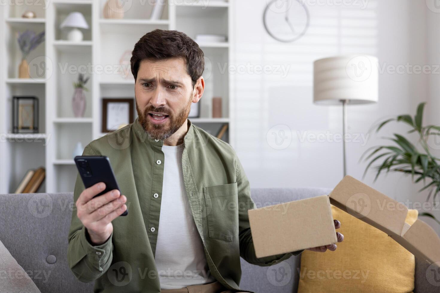 Man looking at phone with shocked expression while holding empty package box. Concept of online shopping disappointment, unexpected delivery issues, or receiving wrong items photo