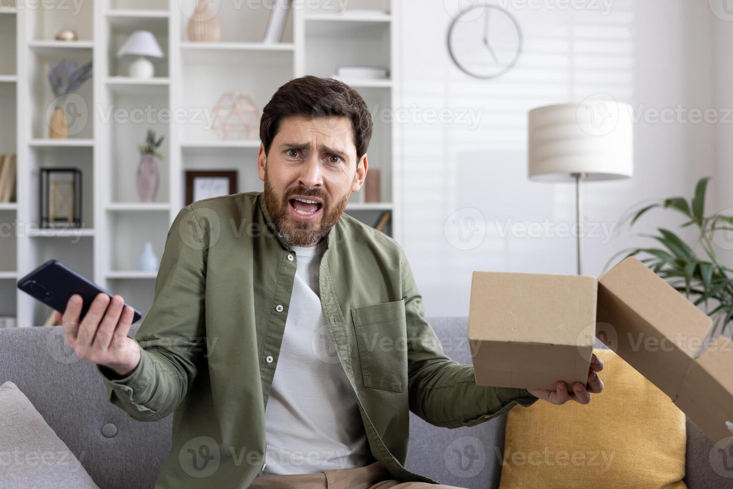 Frustrated man holding smartphone and open delivery box expressing disappointment. Emotional reaction to unsatisfactory online shopping experience, customer dissatisfaction delivery problem at home. photo