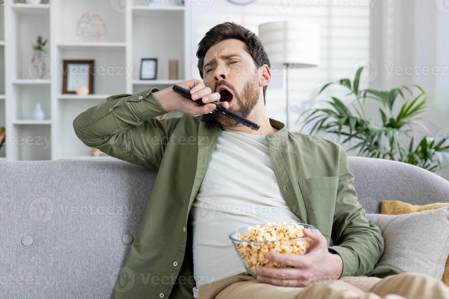 A bored young man yawning while holding a TV remote and a bowl of popcorn, sitting on a sofa in a living room setting. photo