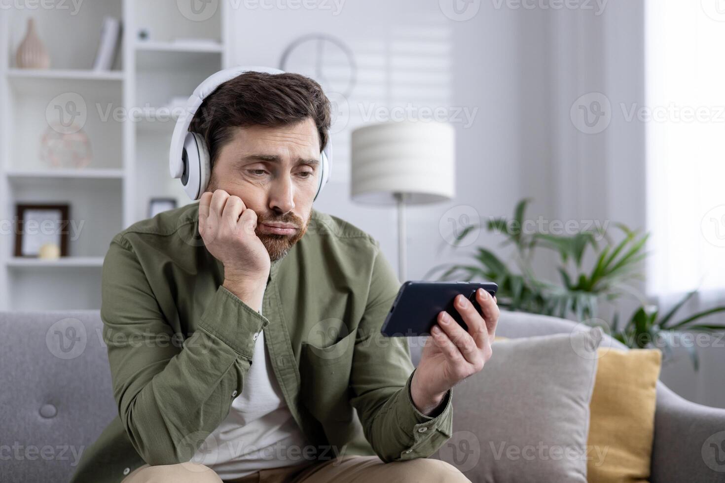Young man sits on couch looking bored while using phone, wearing headphones. Concept of boredom, technology, relaxation, and indoor lifestyle. Captures everyday moments of using modern devices. photo