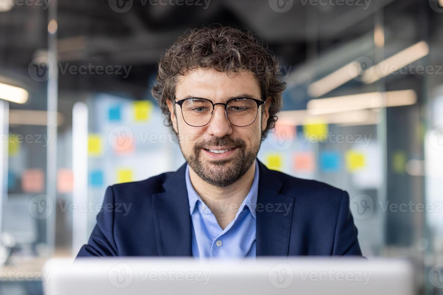 Smiling businessman working on laptop in modern office setting with glass walls and sticky notes. Professional wearing glasses focusing on computer screen photo