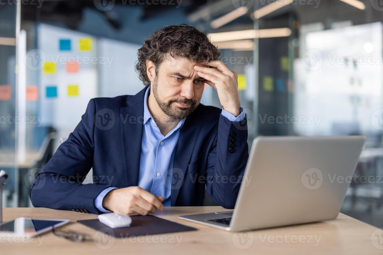 Businessman experiencing stress working on laptop in office. Professional looks frustrated and worried, dealing with issue. Man in suit facing pressure at work. Implements phone and tablet on desk. photo