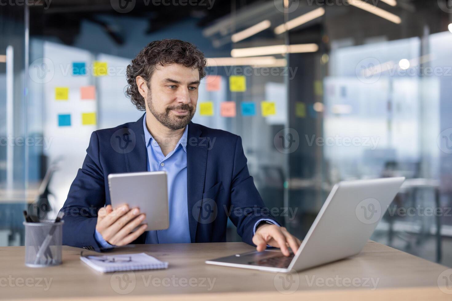 Businessman in formal attire working on laptop and holding tablet in modern office. Corporate workspace with sticky notes and glass walls. Professional environment with focused employee multitasking photo