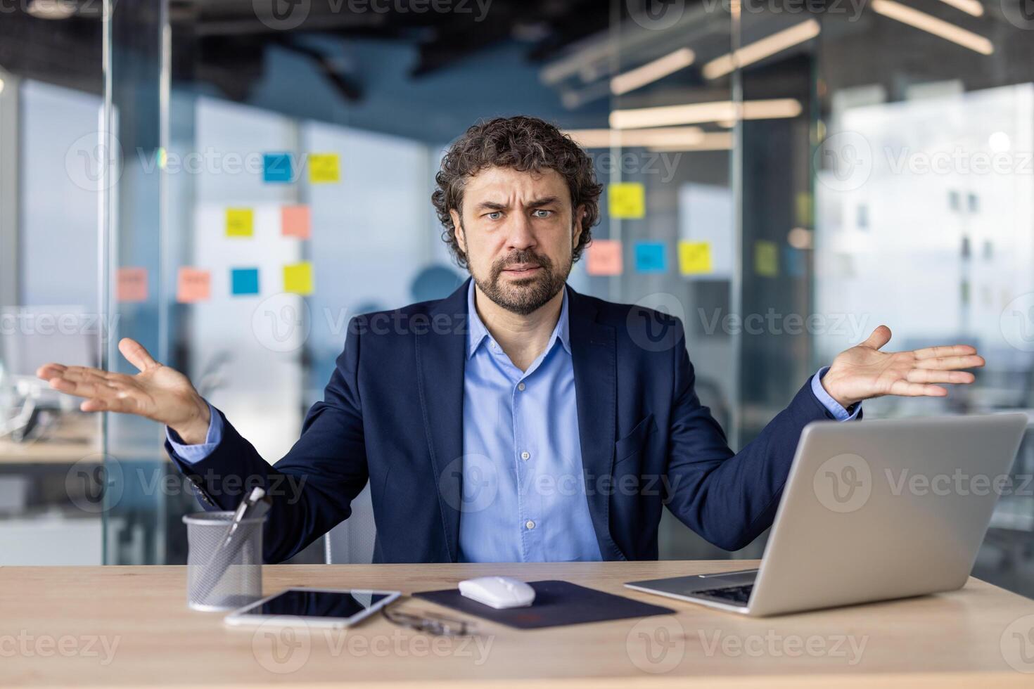 Confused businessman sitting at desk in modern office, shrugging with both hands. Man in suit with laptop, tablet, mouse, and stationery, expressing uncertainty or confusion. photo