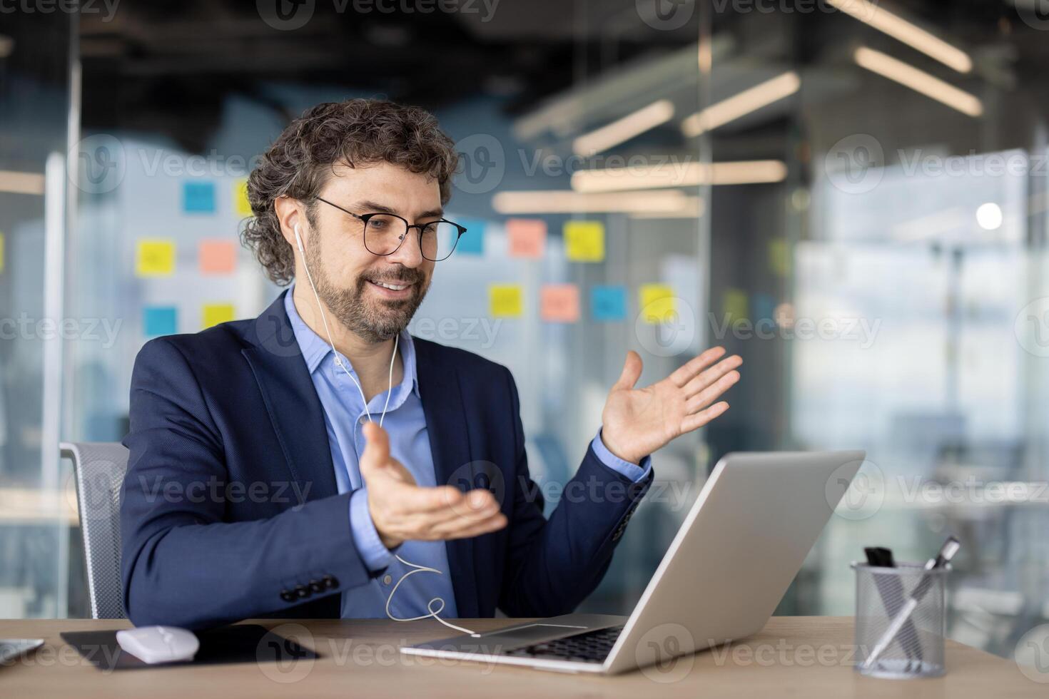 Businessman inside office using laptop for call, man in business suit smiling praying with colleagues at remote online meeting, using headphones. photo