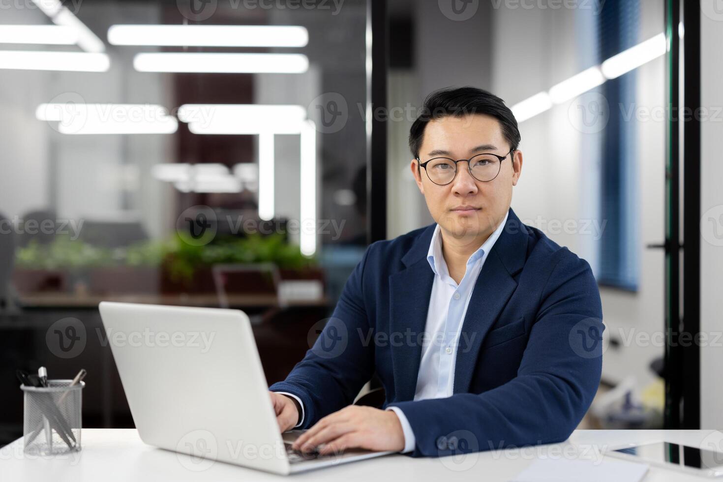 Focused Asian businessman in a suit at his desk in a contemporary office, working intently on a laptop computer. photo