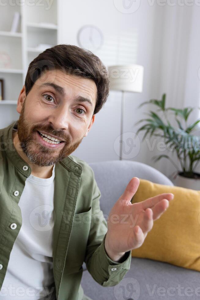 Friendly bearded adult male sitting on a couch in a cozy living room, invitingly gesturing with a warm smile. photo