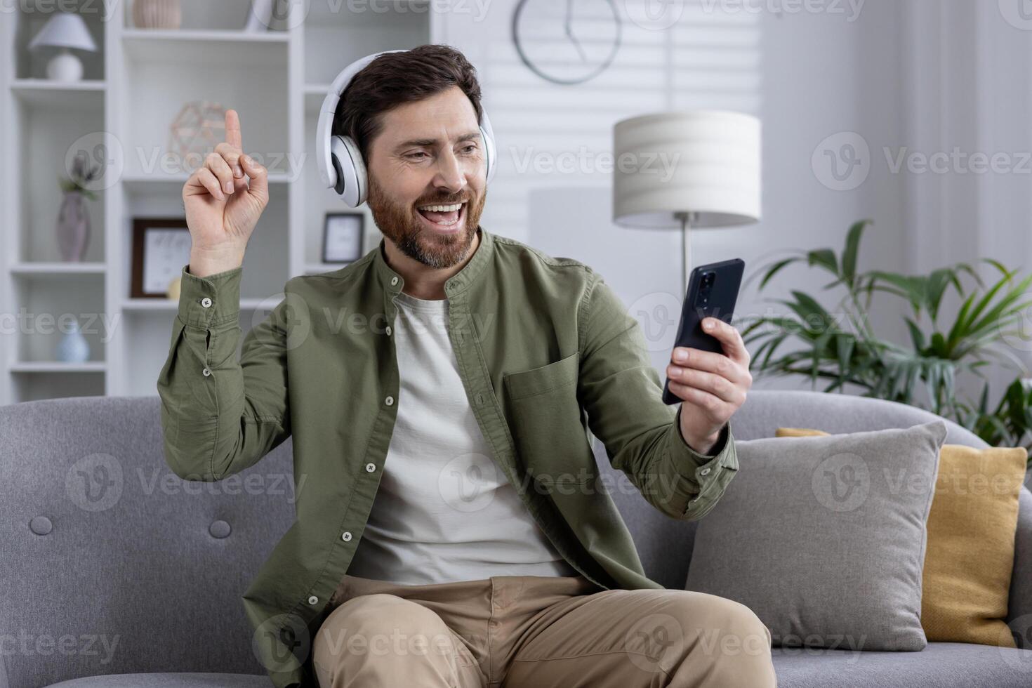 Man wearing headphones holding phone, smiling and pointing while sitting on couch. Enjoying call, music, or online interaction indoors. Casual, relaxed atmosphere capturing positive emotions. photo