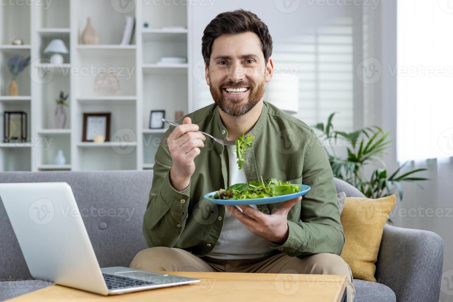 Portrait of a smiling young man sitting at home on the couch in front of the table with a notebook, holding a plate with vegetables and fresh salad, confidently looking at the camera. photo