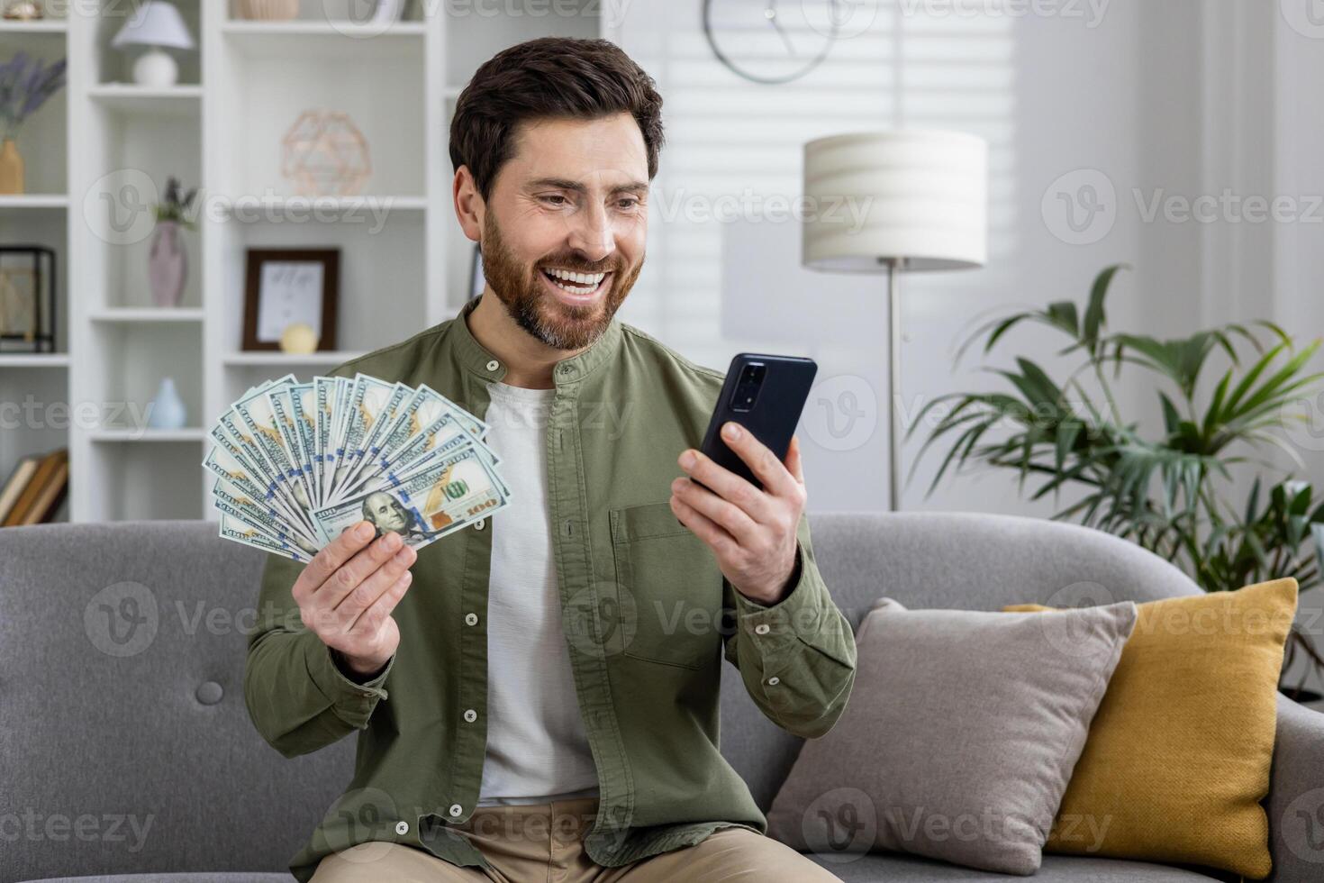 Smiling man sitting on a couch, displaying a spread of dollar bills while happily using his mobile phone at home. photo