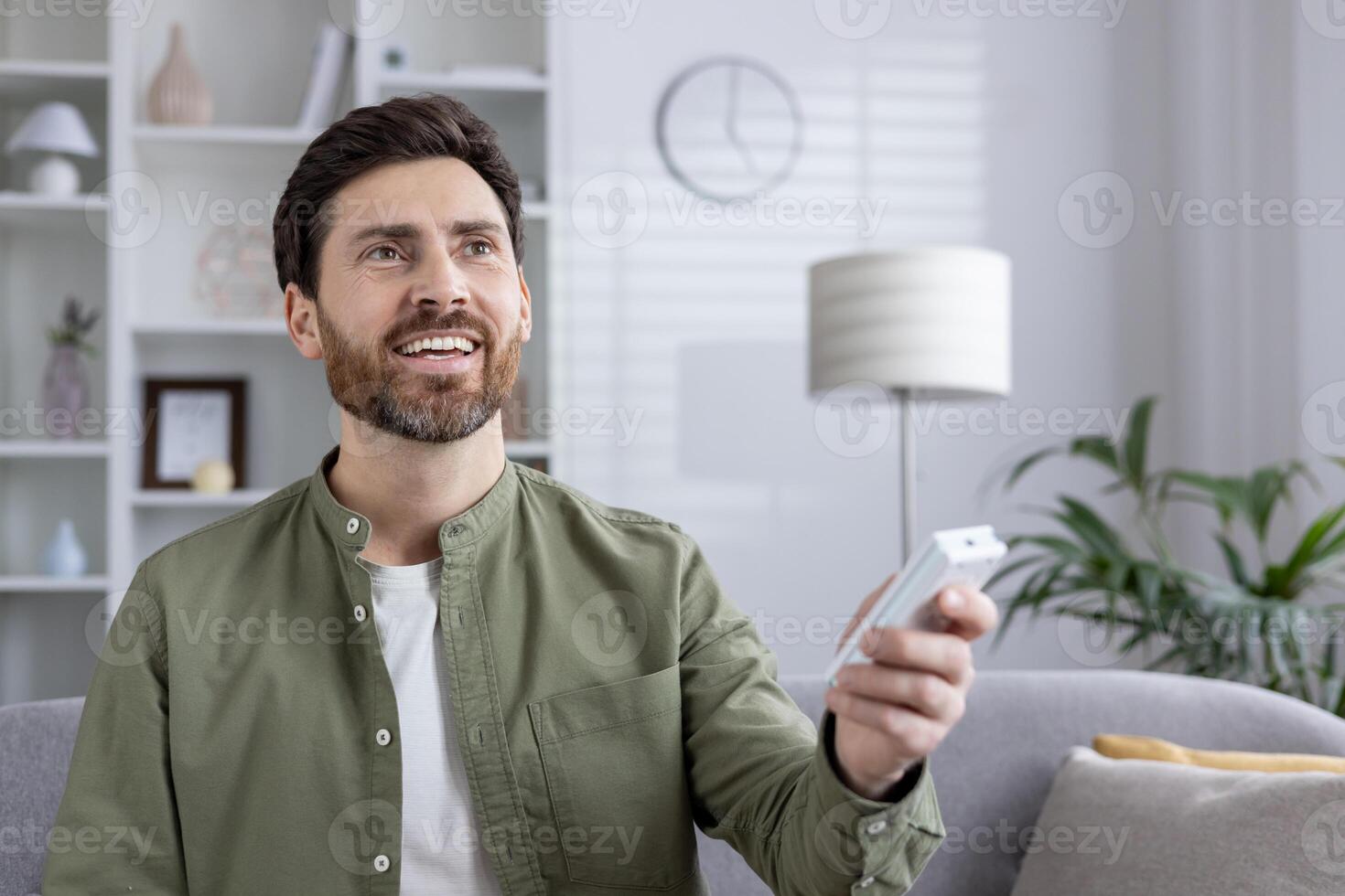 Satisfied man holding air conditioner remote control in hands, smiling indoors. Man adjusting temperature with remote. Concept of comfort, technology, and wellbeing. photo