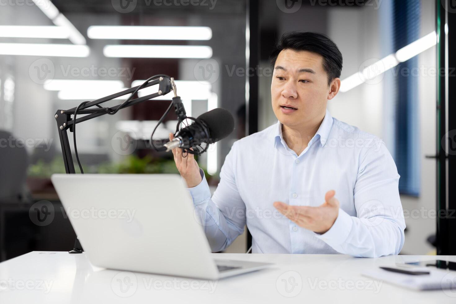 Concentrated korean male recording podcast with professional microphone while looking at laptop screen. Focused financial blogger making new informative content for web site with investment options. photo