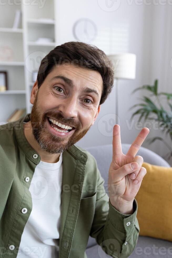 Portrait of happy bearded man making peace sign with two fingers over apartment interior background. Handsome guy taking selfie on smartphone front camera or starting call and greeting friend. photo