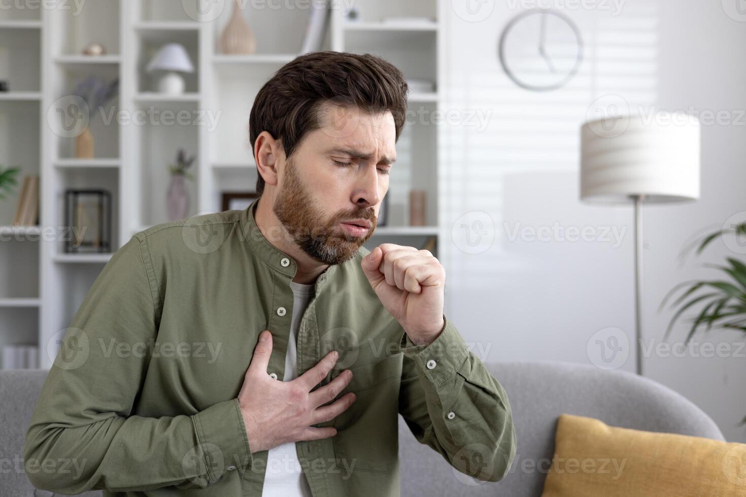 Man with beard, sitting on couch, experiencing chest pain and coughing. Potential health issues like heart problems or respiratory conditions shown. Casual home setting, daytime environment. photo