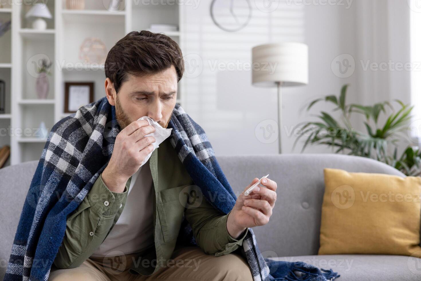 Man suffering from flu sitting on sofa wrapped in blanket holding tissue and thermometer. Expression of discomfort, feeling unwell, checking temperature, and cold symptoms. health and illness concept. photo