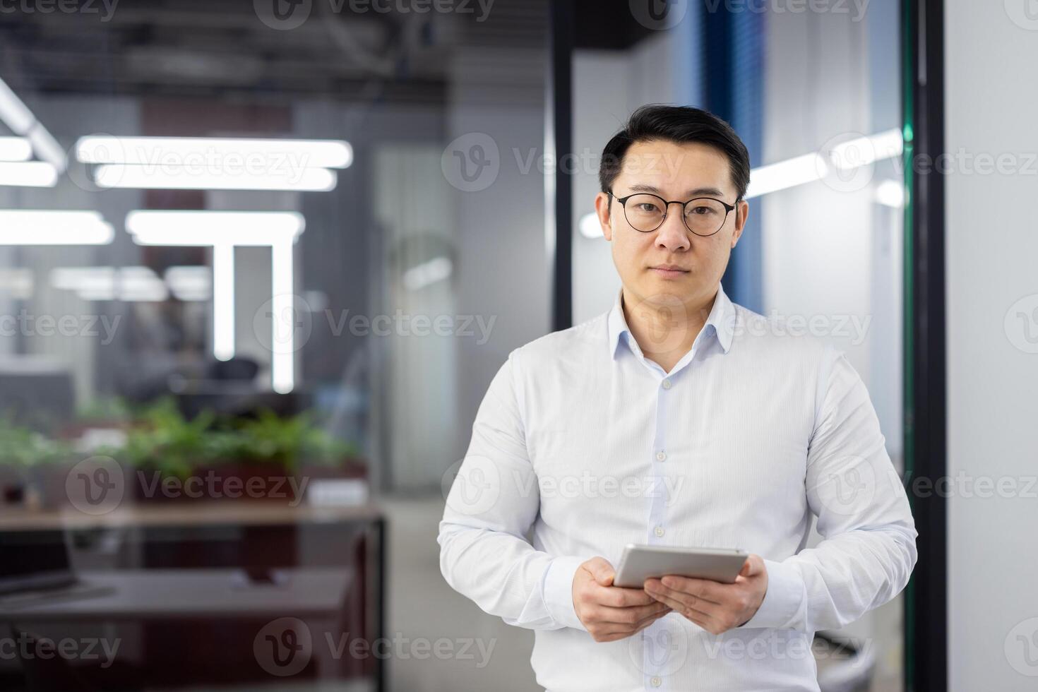 Portrait of a serious and confident Asian man standing in the office in a white shirt and glasses, holding a tablet and looking at the camera. photo