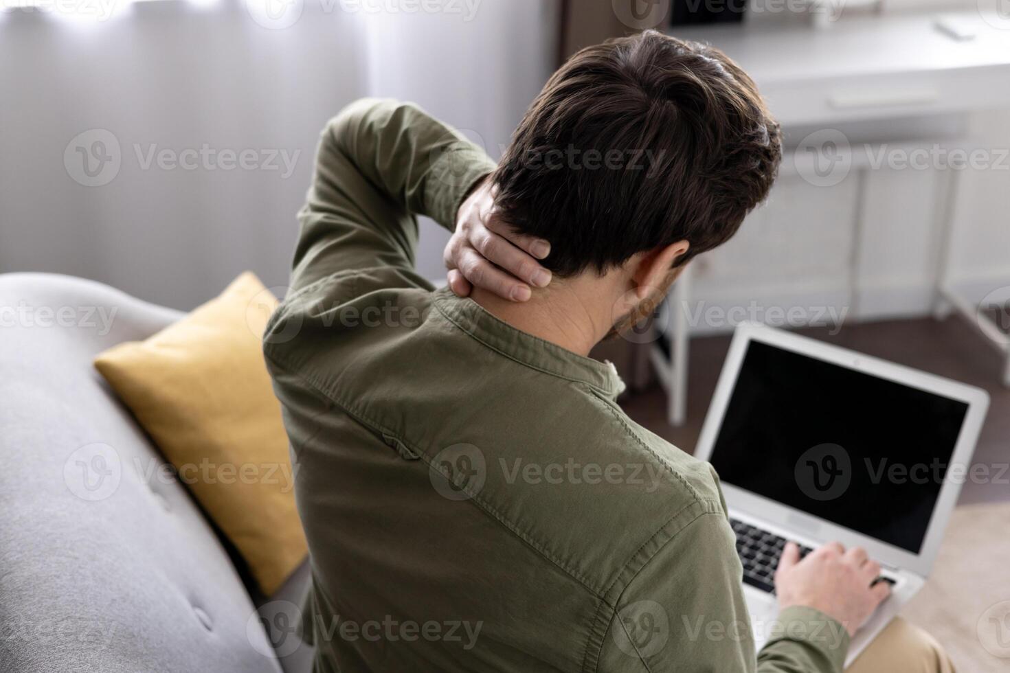 Man experiencing neck pain while using laptop at home. Discomfort from prolonged sitting and computer use. Concept of workplace ergonomics, health, and stress from technology use. photo