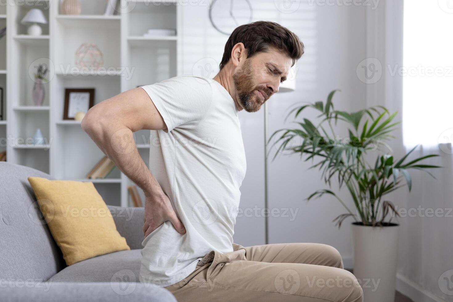 Man sitting on couch holding lower back, experiencing back pain, discomfort or injury. Concept of health issues, chronic pain, posture problems, or muscle strain. photo
