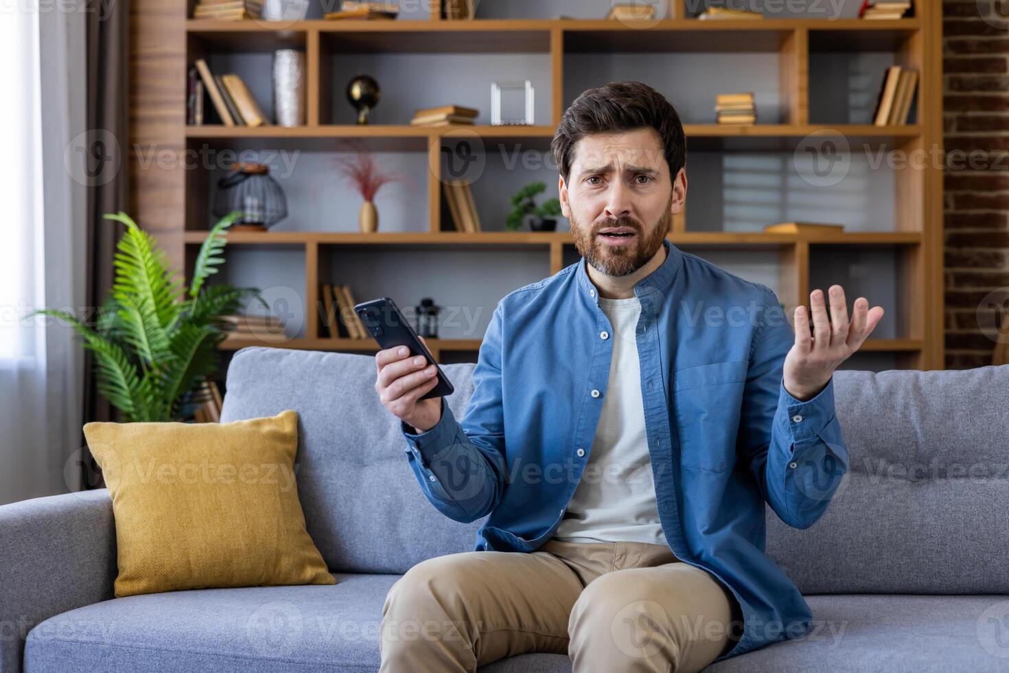 Perplexed adult male sitting on a couch holds a smartphone, expressing confusion and disbelief in a home environment. photo
