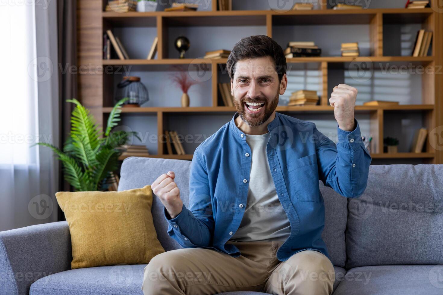A joyful moment captured of a man seated on a gray sofa in a cozy living room, raising his fist in a triumphant gesture of victory or success. photo