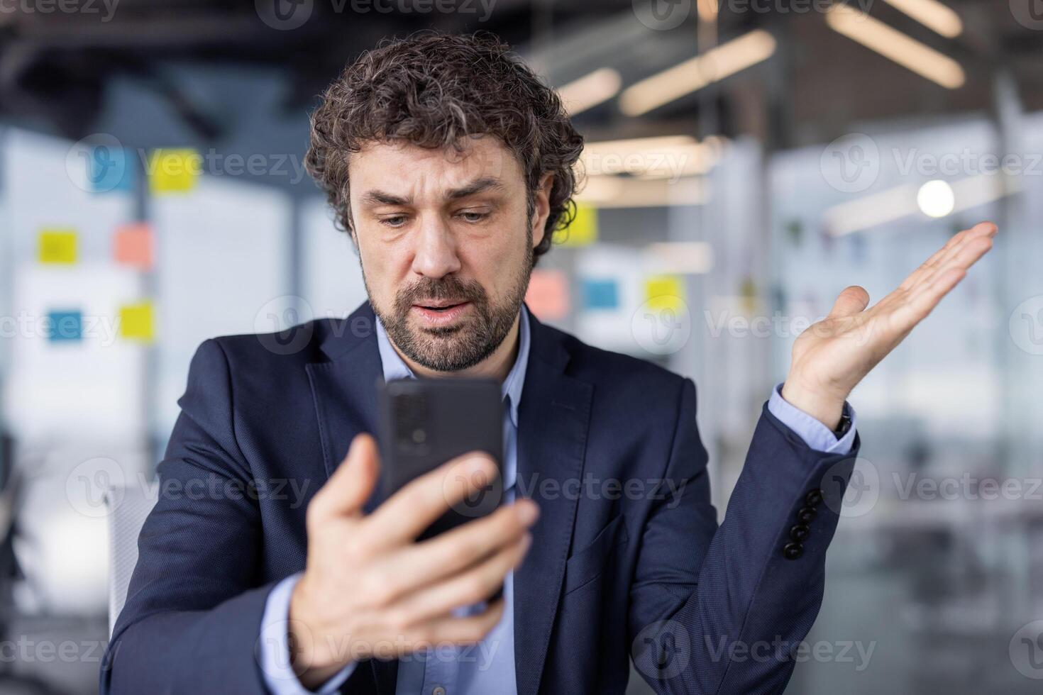 Confused businessman in suit looking at cell phone in modern office setting. Man appears perplexed or frustrated while holding smartphone, likely dealing with unexpected news or information. photo