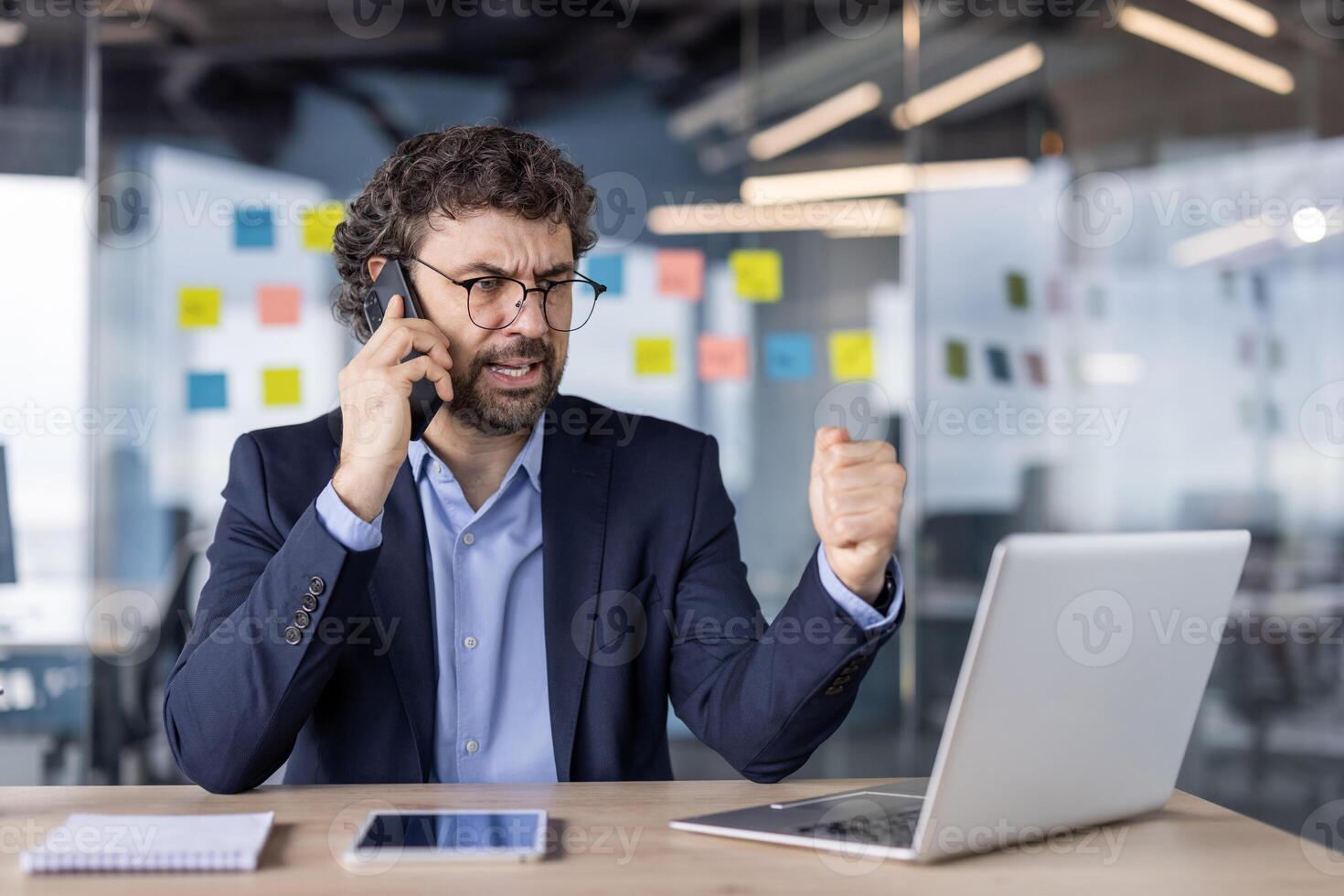 Businessman showing frustration during phone call in modern office environment. Man in suit expressing anger while working on laptop, with digital tablet and notebook on desk. photo