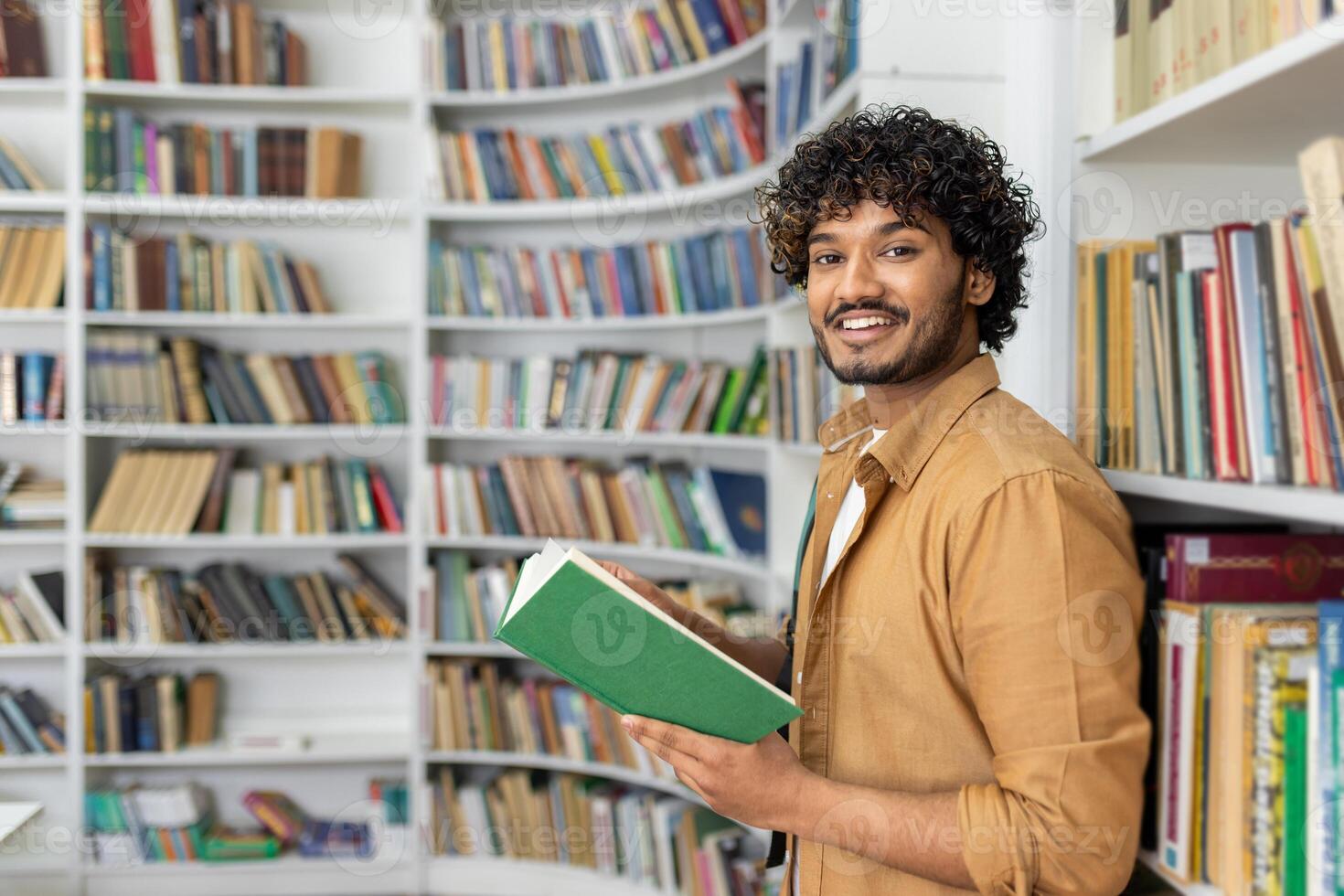A focused man in casual attire reads a green book in a well-stocked, organized library setting, suggestive of lifelong learning and education. photo