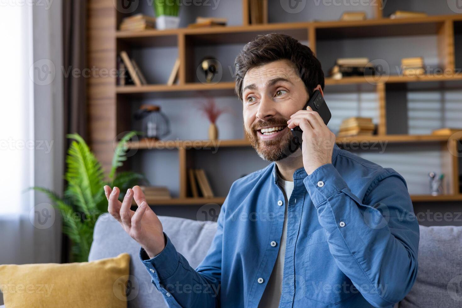 A casually dressed man in a blue shirt gestures as he speaks on a mobile phone in a modern living room setting. photo