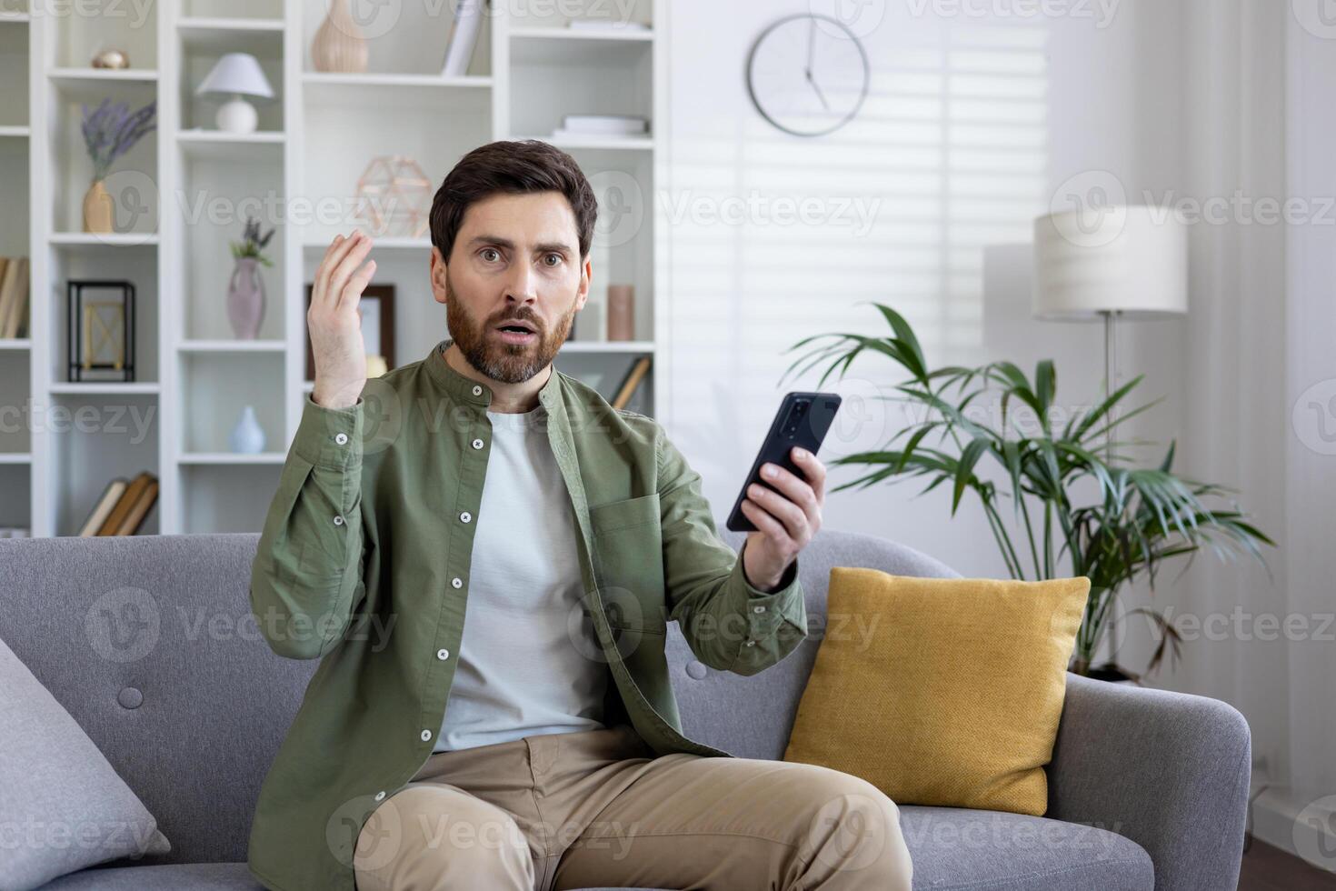 Adult man expressing shock and confusion while looking at his smartphone, seated comfortably on a living room sofa. photo