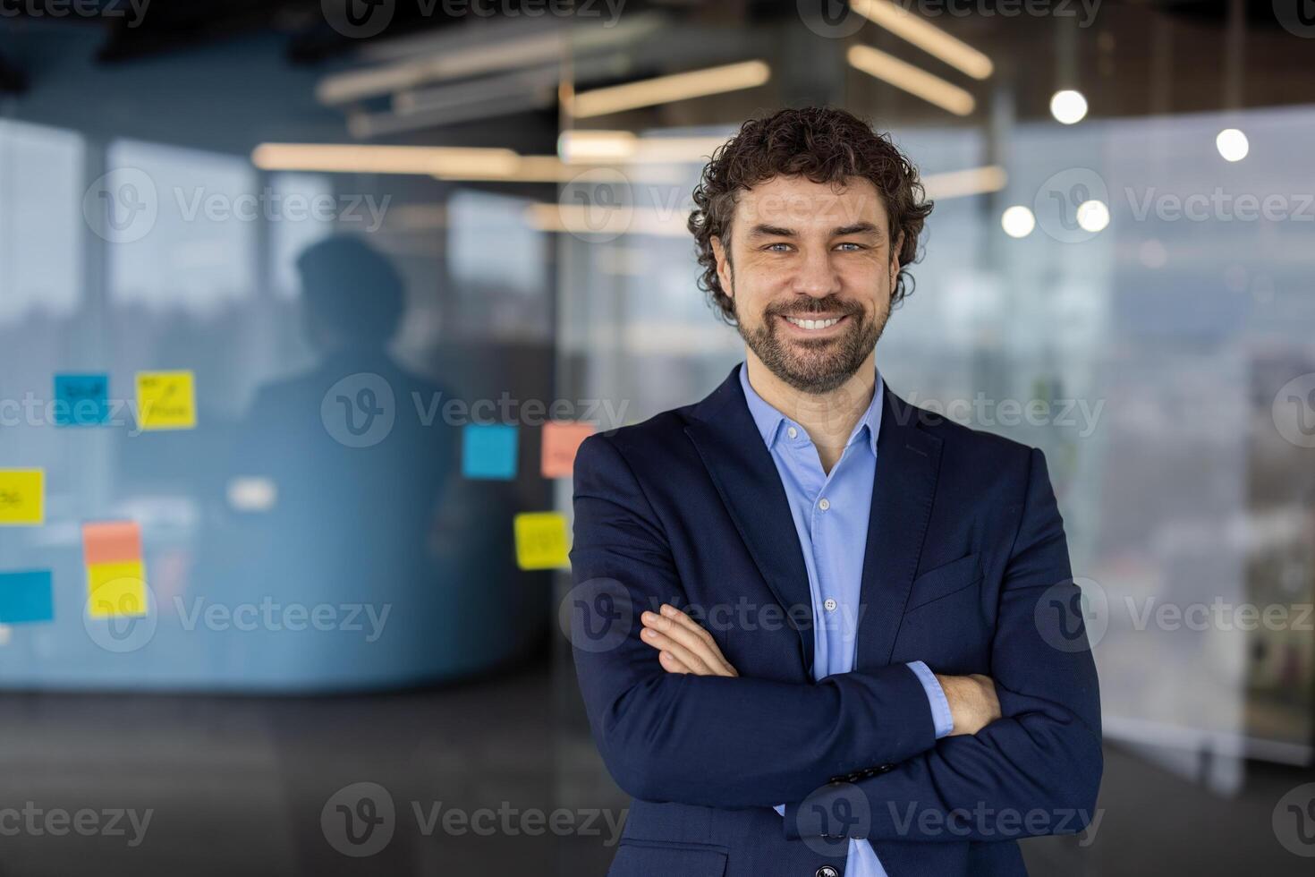 Portrait of successful mature boss, senior businessman in glasses and business suit looking at camera and smiling, man with crossed arms working inside modern office building photo