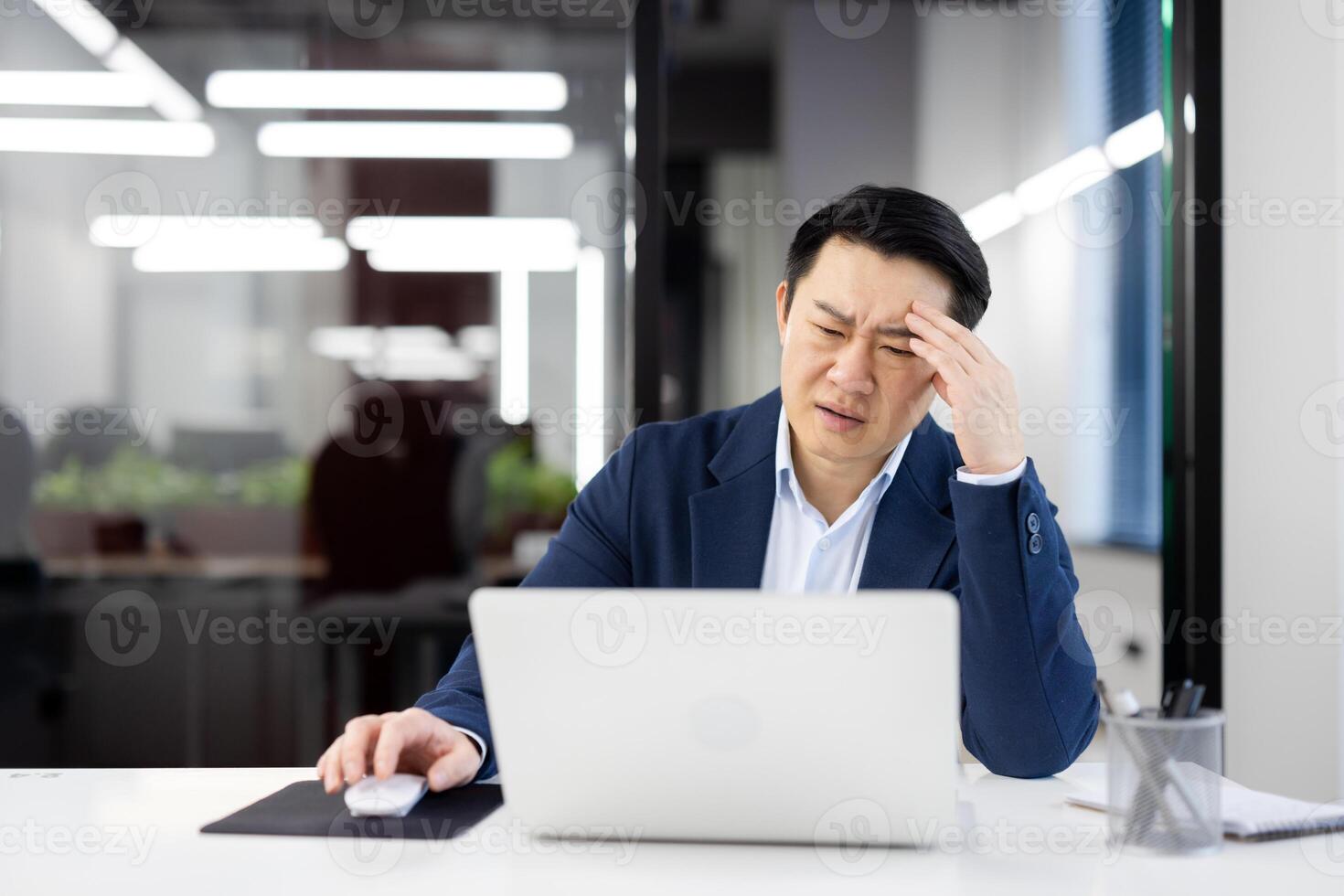Frustrated Asian businessman experiencing stress and headache while working on a laptop in a modern office environment. photo