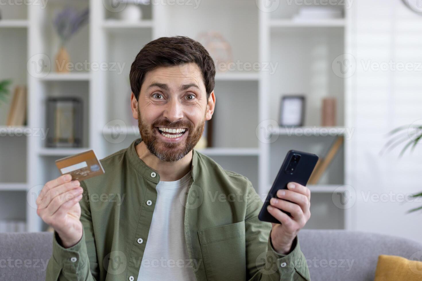 Excited buyer with debit card and mobile phone in pleasant shock from online shopping from home. Amazed male customer getting massive cash back and extra bonuses after making order with shipping. photo